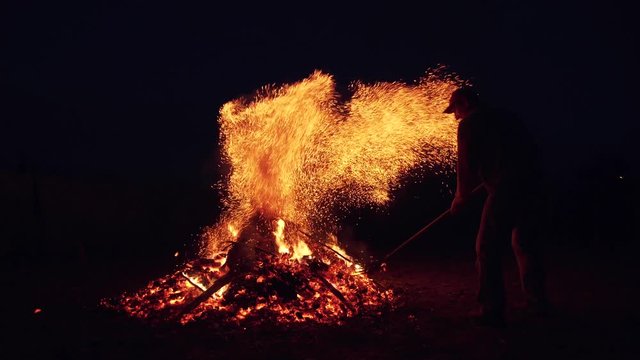 a man straightens forks burning coals in the fire. sparks of a fire against the dark sky