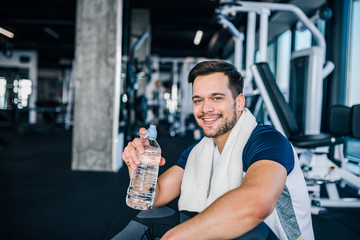 Fototapeta na wymiar Resting time. Smiling young man in sportswear sitting on the floor with bottle of water and looking at camera.