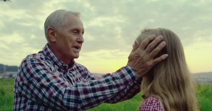 Portrait shot of the caucasian old grandfather stroking his teenage cute granddaughter while they standing in the field.