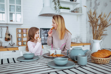 Happy little girl and her beautiful young mother have breakfast together in a white kitchen. They drink milk and eat cookies. Maternal care and love. Horizontal photo