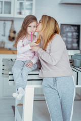 Obraz na płótnie Canvas Happy little girl and her beautiful young mother have breakfast together in a white kitchen. Mom hugs and kisses daughter. Maternal care and love. Vertical photo