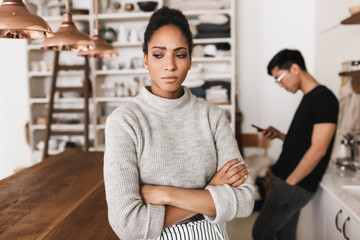 Upset african american woman holding hands together sadly looking aside with asian man holding cellphone on background. Young international couple in quarrel spending time on kitchen