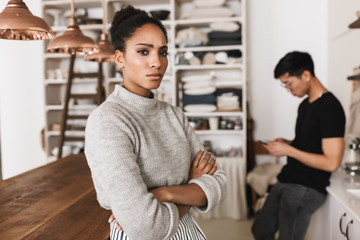 Upset african american woman holding hands together sadly looking in camera with asian man holding cellphone on background. Young international couple in quarrel spending time on kitchen