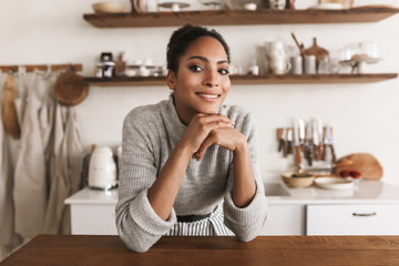 Young pretty smiling african american woman with dark curly hair leaning on table happily looking aside while spending time in beautiful cozy kitchen at home