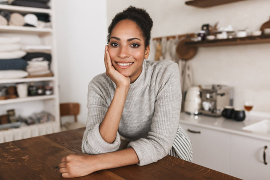 Young Attractive Smiling African American Woman With Dark Curly Hair Leaning On Table Happily Looking In Camera While Spending Time In Beautiful Cozy Kitchen At Home