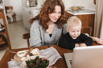 Young smiling woman with red hair in knitted sweater and little son sitting at the table with food and watching cartoons on laptop. Mom happily spending time with her baby in kitchen at home