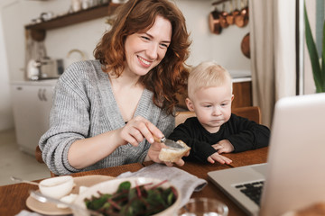 Young cheerful woman with red hair in knitted sweater and little son sitting at the table with food and happily watching cartoons on laptop. Mom spending time with her baby in kitchen at home