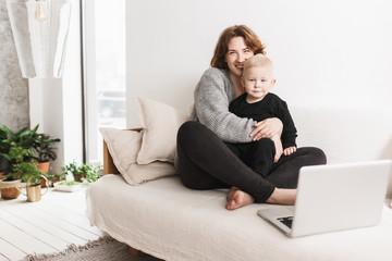 Obraz premium Young smiling woman sitting on sofa with her little beautiful son happily looking in camera. Mom and baby boy joyfully spending time with laptop in living room at home