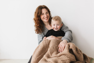 Young beautiful smiling mom with red hair in knitted sweater sitting on floor with her little son...