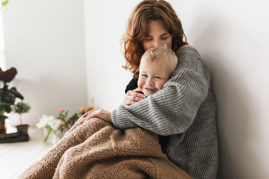 Young beautiful mom with red hair in knitted sweater sitting on floor kissing her little son covering with blanket happily spending time together at home