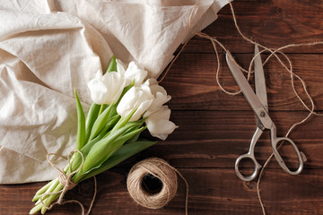 Spring bouquet of white tulip flowers, blank paper card, scissors, twine on rustic wooden desk. Womens day composition on flat lay style, above view.