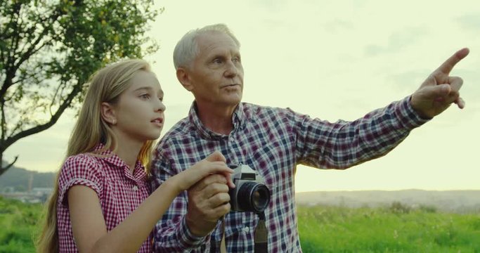 Portrait shot of the gray haired old grandpa telling and showing to his pretty teenage granddaughter to take photos with the camera in the field.