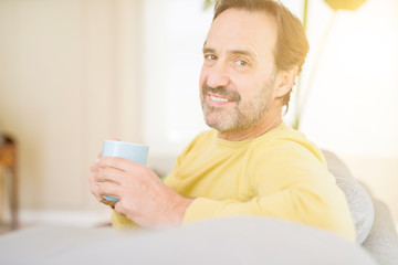 Handsome middle age man sitting on the sofa relaxed drinking a cup of coffe and smiling at the camera at home
