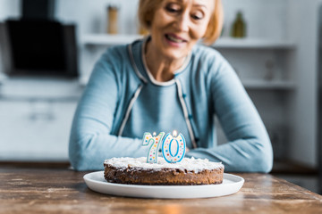 selective focus of cake with '70' sign on top and smiling senior woman on background during...