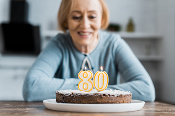 smiling senior woman sitting at table and looking at cake with '80' sign on top during birthday...