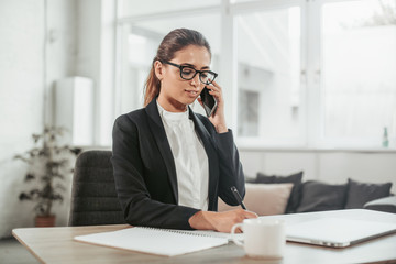 A picture of nice brunette sits at table and talks on the phone. She writes down information. Girl wears glasses and costume