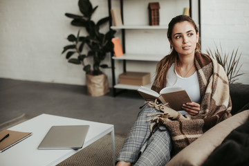 Attractive young woman sits on sofa and looks to the right. She holds book in hands. Girl has brown...