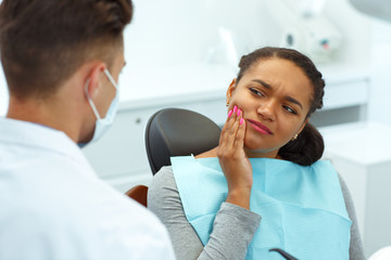 Young woman having toothache, covering mouth by hand and shrugged from pain, sitting in dental chair. Professional dentist wearing in mask and protective glasses, helping patient to cure.