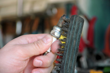 Cleaning a car engine spark plug with wire brush in car garage. Mechanic is brushing used spark plug with a stainless steel brush in auto mechanics workshop. Worker with service tools in hand.