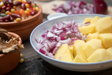 Sliced potato and onion on ceramic plate, and other vegetables, spices and dried mushrooms in the background. Old wooden table.