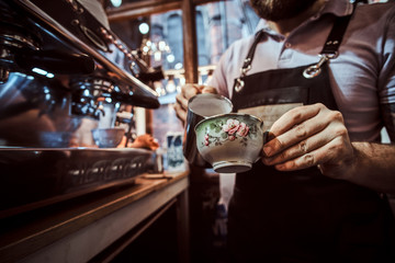 Barista in apron making a cappuccino, pouring milk in a cup in a restaurant or coffee shop