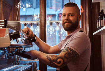 Cheerful barista with stylish beard and hairstyle making coffee for a customer in the coffee shop  - obrazy, fototapety, plakaty