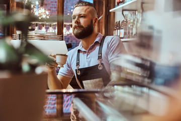 Handsome barista in apron drinking coffee during lunch break standing behind the counter in the coffee shop