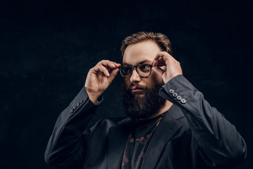Obraz premium Portrait of a smart bearded guy wearing a black jacket corrects his glasses while standing in a dark studio