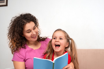 Obraz premium An little daughter and her mother spending time together. They sit on the couch and read a book.