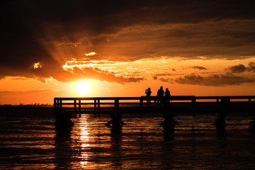 Fototapeta premium Silhouette of young adults on a fishing pier watching the sunset. Hanging out with friends, enjoying nature. Long Island New York. 