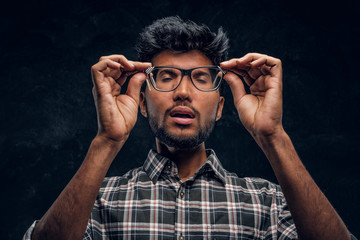 Indian young scholar in altered minds holding glasses and closing his eyes. Studio photo against a dark textured wall