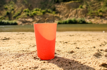 an orange glass stands in the sand, on the shore of the pond