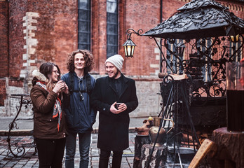 Two guys and a girl choose souvenirs, drink hot coffee, pose and smile at the camera walking through the city square