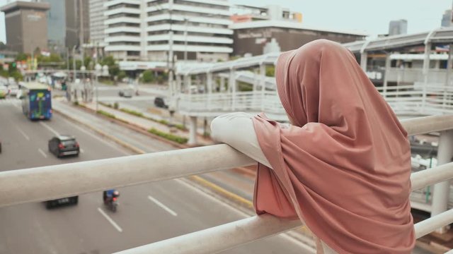A young, sad Muslim girl stands on a bridge over road traffic in downtown Jakarta. Indonesia.