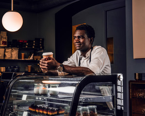 Barista holds a cup of coffee leaning on the counter at the coffee shop and looks at the camera