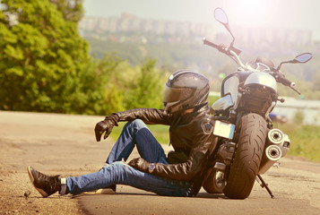 Biker in outfit sits next to motorcycle on asphalt. Motorcyclist rests near the motorbike. Toned.