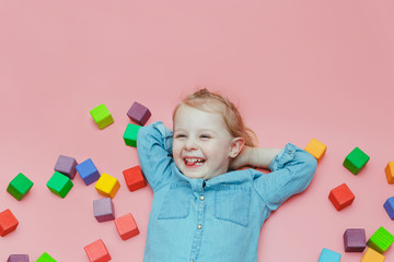 A charming little girl in denim clothes is lying on a pink background among wooden colored cubes...