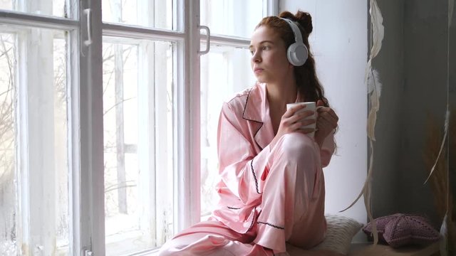 Young Female Listening To Music In Headphones Sitting By The Window