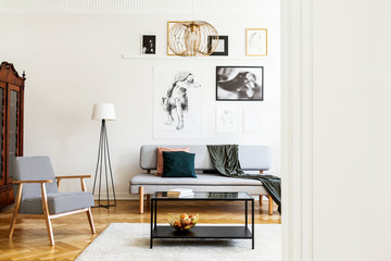 Table on carpet next to armchair in living room interior with posters above grey couch. Real photo