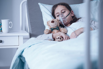 Sad kid with cystic fibrosis lying in a hospital bed with oxygen mask and plush toy