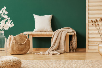 Wooden bench with beige carpet and pillow next to straw handbag, copy space on the empty green wall