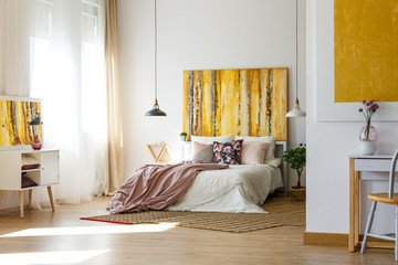 Floral pillow and pastel pink bedding on king size bed in chick bedroom interior with abstract painting on the wall and checkered carpet on wooden floor