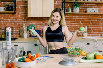 Young smiling woman holding broccoli and green apple while standing in the kitchen. Healthy Food. Dieting Concept. Healthy Lifestyle. Cooking At Home.