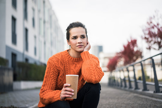 Portrait of pensive woman with coffee to go wearing orange knit pullover outdoors