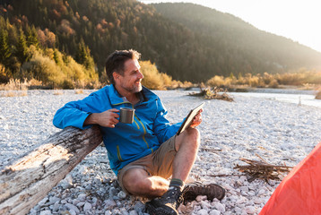 Mature man camping at riverside, using tablet