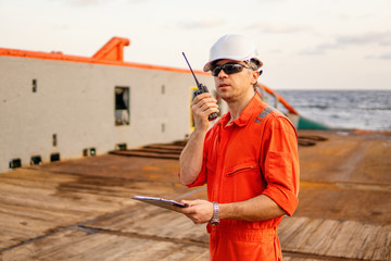 Marine Deck Officer or Chief mate on deck of offshore vessel or ship , wearing PPE personal protective equipment - helmet, coverall. He speaks VHF walkie-talkie radio in hands.