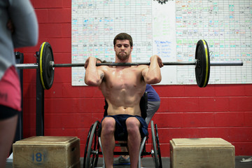 Shirtless male adaptive athlete lifting barbell while sitting on wheelchair against wall in gym