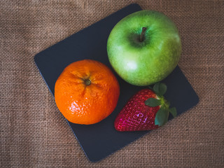 A plate of black slate with a green apple, a tangerine and a strawberry on a burlap background in vintage style