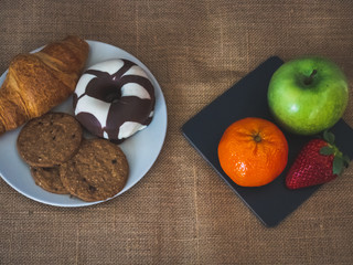 A plate of black slate with a green apple, a tangerine and a strawberry and one plate with one croissant, one chocolate donut and cookies on a burlap background in vintage style
