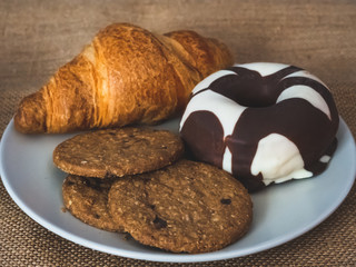 One plate with one croissant, one chocolate donut and cookies on a vintage burlap background